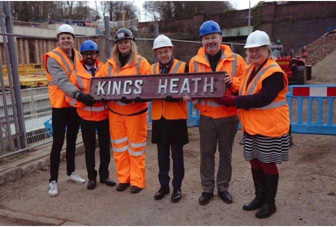 Five people in high visibility jackets hold up an old station platform sign saying "Kings Heath"